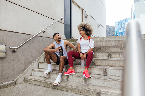 A shot of two friends, one male and one female, taking a break together after a gym session in Newcastle Upon Tyne, North East England. They are both smiling, dressed in activewear and holding reusable water bottles.