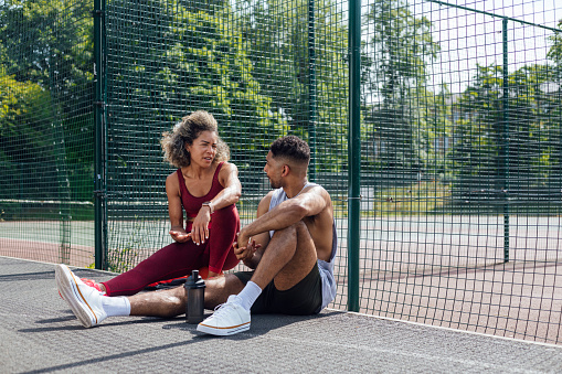 A shot of two friends, one male and one female, taking a break together on a basketball court in a park in Newcastle Upon Tyne, North East England. They are both wearing activewear, looking each other and talking.