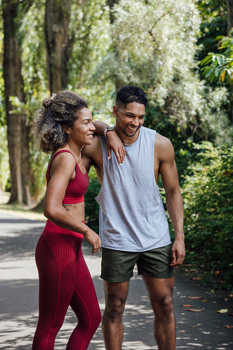 A three-quarter-length portrait of two friends, one male and one female, standing and laughing together in a park in Newcastle Upon Tyne, North East England. They are both wearing activewear and the female has her arm resting on her friend's shoulder.