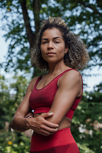 A three-quarter-length portrait of a multiracial female with a natural green backdrop in a park in Newcastle Upon Tyne, North East England. She is smiling, has one hand on her hip and is wearing activewear.