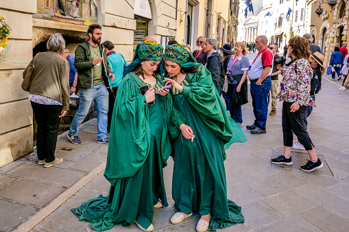 Assisi, Umbria, Italy, May 04 -- Two women in medieval costume take a break during the celebrations of Calendimaggio (May Day Festival) in the historic heart of Assisi, in Umbria, with some tourists flocking to the small town. Celebrated by the ancient Romans as an ode to spring, this ancient festival coincides with the Roman Kalends which paid homage to the blossoming of trees and flowers and the beginning of the good season. During the Calendimaggio all the medieval districts of Assisi, divided into Parte di Sopra and Parte di Sotto, challenge each other in skill competitions with the crossbow and the rope, in singing competitions and allegorical parades. Famous for being the city of San Francesco and Santa Chiara (St Francis and St Clare), Assisi is recognized in the world as a place of spirituality and peace between all peoples and different confessions. Since 2000 the Franciscan sites of Assisi have been declared a World Heritage Site by UNESCO. Wide angle mage in high definition quality.