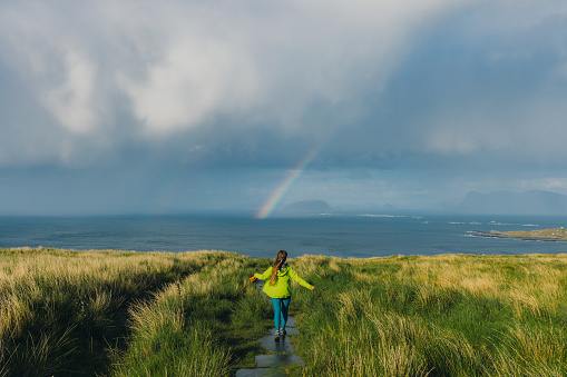 Happy female contemplating the trip on beautiful island with bright rainbow in the sky in Scandinavia