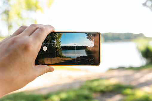 A smartphone with a photo of a lake held by a male hand against the background of a lake surrounded by forest on a sunny day.