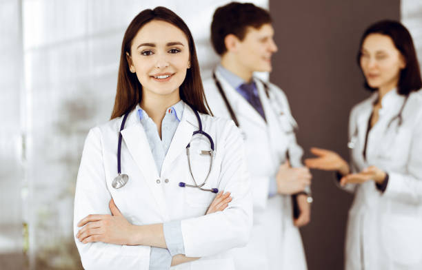 une femme-médecin intelligente et souriante se tient debout, les bras croisés, dans une clinique, avec ses collègues à l’arrière-plan. portrait de médecins au travail. service médical parfait dans un hôpital - portrait doctor paramedic professional occupation photos et images de collection