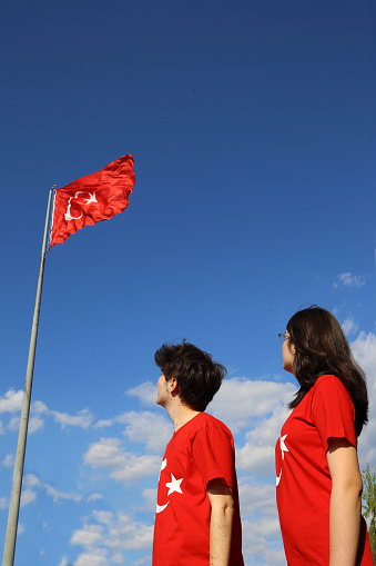 Two young people in red t-shirts looking at the turkish flag in front of the blue sky view.