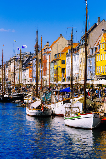 People and tourists enjoying themselves at the harbor in the Nyhavn area of Copenhagen, Denmark. Boats are moored up on the water, while beyond are the colorful facades of bars and restaurants.
