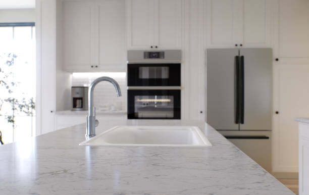 istock Large white kitchen with island and all necessary appliances. 3d rendering. Emphasis on a stone top with white marble, in the background is an oven, a sink from a mixer and a refrigerator. 1487779080