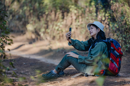 Woman is resting in the forest and holding mobile phone, The climber rests. Hikers with backpacks sitdown resting in forest. Traveler tired and sit down in the forest with backpacks.