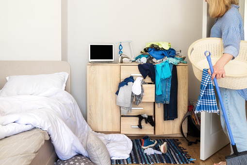 General plan of the bedroom. A woman came into her son's room with cleaning products to clean up the mess. She holds a basin in her hands to collect dirty laundry for washing and clean the apartment.