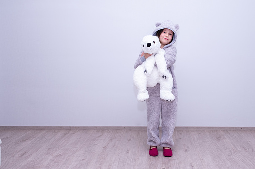 Portrait of a Caucasian baby girl 7 years old in a plush overalls hugs a soft toy, a polar bear. The child stands against the background of a gray wall. Place for text.