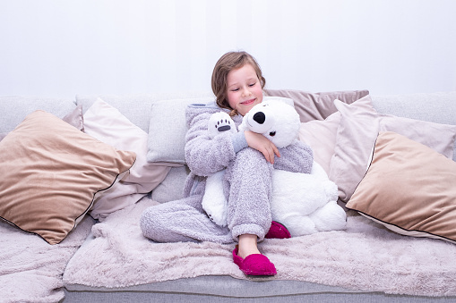 A portrait of a Caucasian child of a girl of 7 years old in a plush overalls sits on a large soft sofa and hugs a soft toy, a polar bear.
