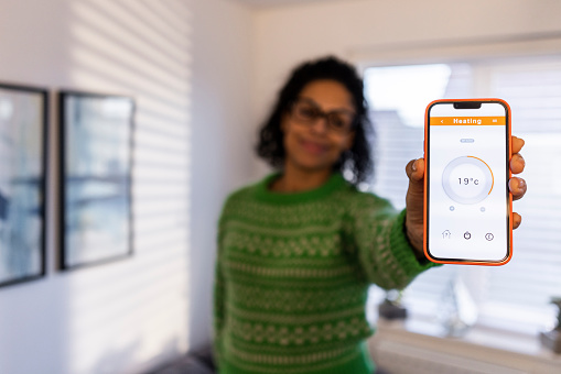 A wide angle shot of a multicultural woman standing in her living room at home as she holds out her mobile phone towards the camera. She is showing a smart metre app on the phone screen. She is out of focus in the background.