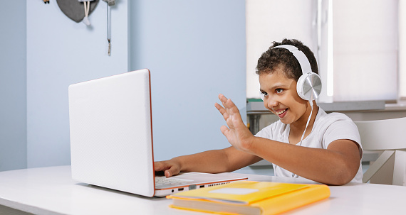 A Black boy in a yellow T shirt works on a laptop and communicates via video conference with a teacher. Children studying from home