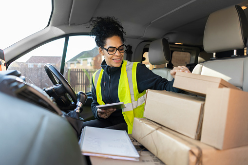 A shot of a multicultural female postal worker wearing a high visibility vest about to make a delivery in an urban housing estate. She is smiling as she organises her parcels. She is holding a digital tablet in her hand as she does so.