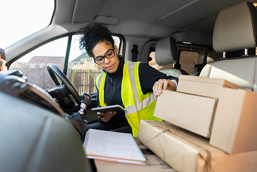 A shot of a multicultural female postal worker wearing a high visibility vest about to make a delivery in an urban housing estate. She is organising her parcels before deliving them. She is holding a digital tablet in her hand as she does so.
