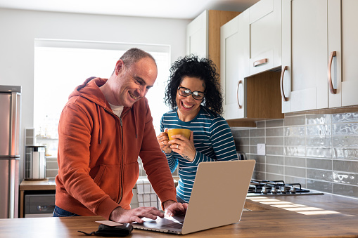 A shot of a mature heterosexual couple standing in their kitchen at home. They are looking at their computer together and making plans for the future. The woman is holding a hot beverage in her hand. They are both smiling and showing positive emotion.