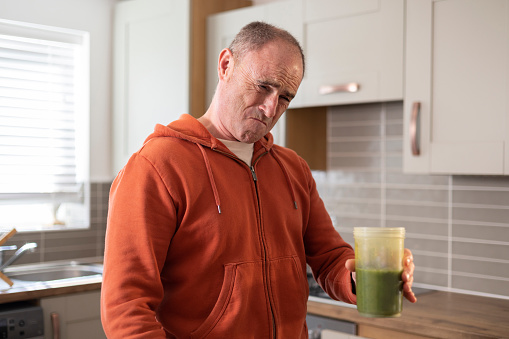 A waist up shot of a mature male standing in his kitchen at home. He is tasting homemade vegetable juice that he has prepared. He is pulling a bitter facial expression.