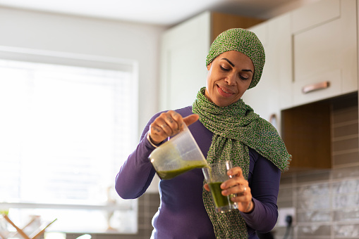 A waist up shot of a multicultural female pouring a green smoothie into a glass in her kitchen at home. She is wearing a green head band and scarf around her neck. She Is smiling and portraying positive emotion.