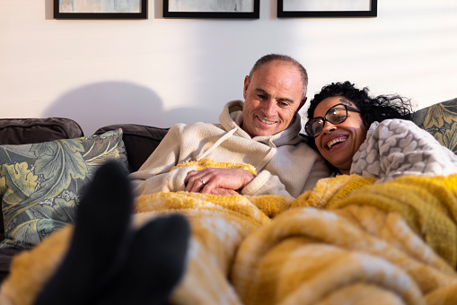 A shot of a mature heterosexual couple laying on the sofa together with a blanket over them. They are staying warm during the cost of living crisis.