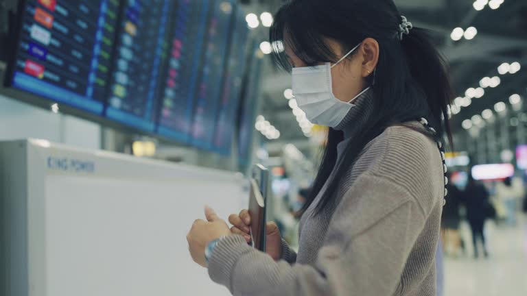 Asian woman wear surgical mask, using smart phone and watch wristwatch for check Departure Board timetable at the airport