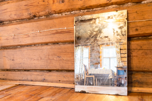 windows, table and chairs of village house are reflected in old mirror standing on floor of log cabin (focus on the reflection in the mirror)