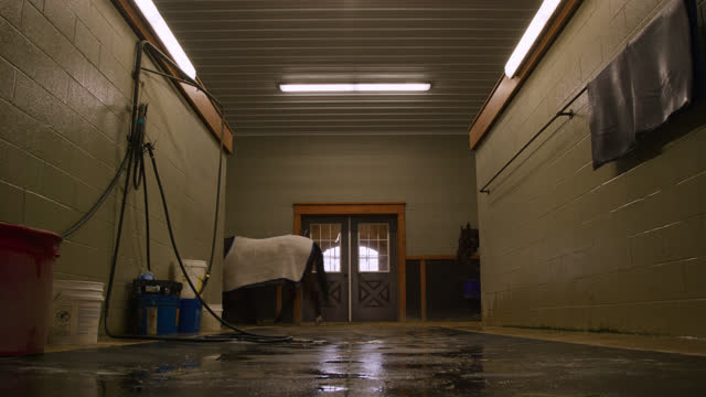 Long Shot of Thoroughbred Racehorses and Their Handlers Walking Past a Wash Station in a Barn