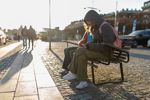 Group of people sitting on the bench on square in Stockholm and resting on they touristic tour. Teenage girl with grey winter jacket with hood tipping messages on mobile phone.