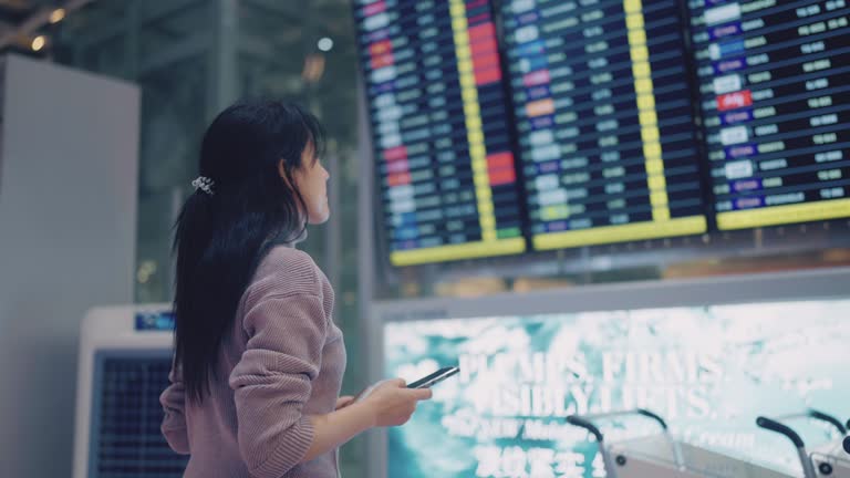 Asian woman using smart phone for checking flight schedule board in airport terminal.