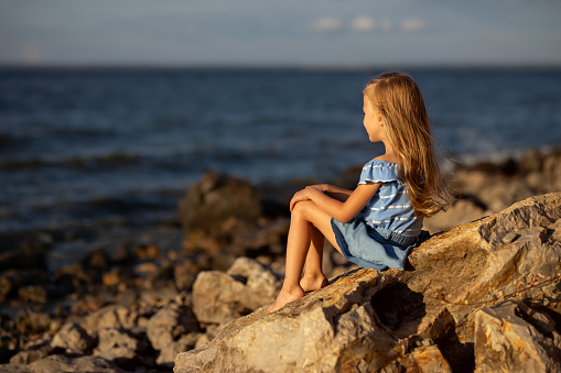 Girl with long blond hair sits on a stone on the banks of a river, lake, sea. She is looking at ocean and thinking dreamily. Girl sitting on rocks. Vacation on sea.