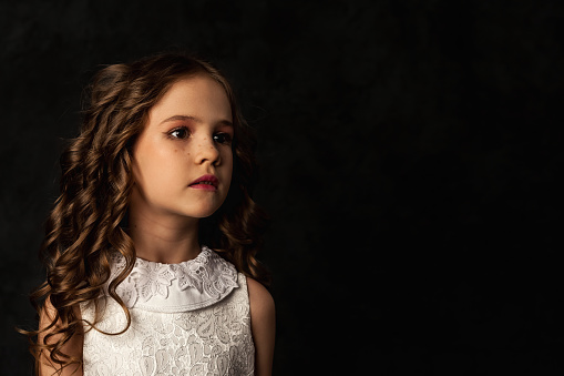 Cover girl with freckles in doll image 6-7 year old isolated in shadow, frozen look pose, looking away. Lovely kid model in white dress at black. Theatrical performance concept. Copy ad text space