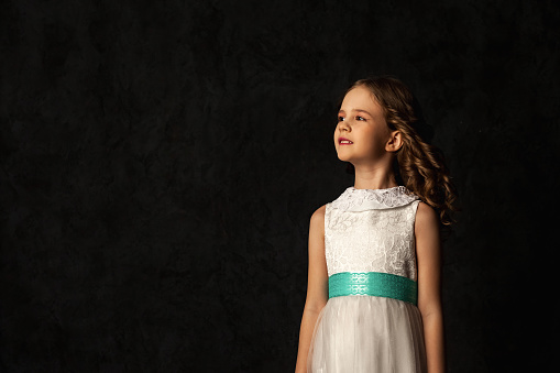 Portrait of little cover girl model with freckles in doll image at black background, looking away. Smiling kid 6-7 year old in white dress isolated in shadow. Fashion style concept. Copy ad text space