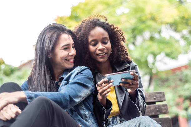 istock Female friends using mobile phone sitting on bench at city 1487834427