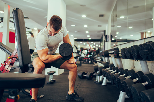 A healthy sportsman is exercising with dumbbells while sitting in a gym. A muscular sportsman in shape is sitting at the sports center and doing exercises for the biceps and triceps with a dumbbell.