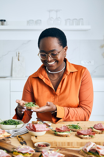 Cheerful young girl preparing sandwiches for social gathering or birthday event, with fresh green salad, ham, cheese, mayo, mustard, tomato, cucumber, pesto, cottage cheese, cream cheese, avocado, Spanish onion with healthy seasoning, herbs and spices, served on a wooden home table, representing a wellbeing and a healthy lifestyle, food indulgence and joy, an image with a copy space