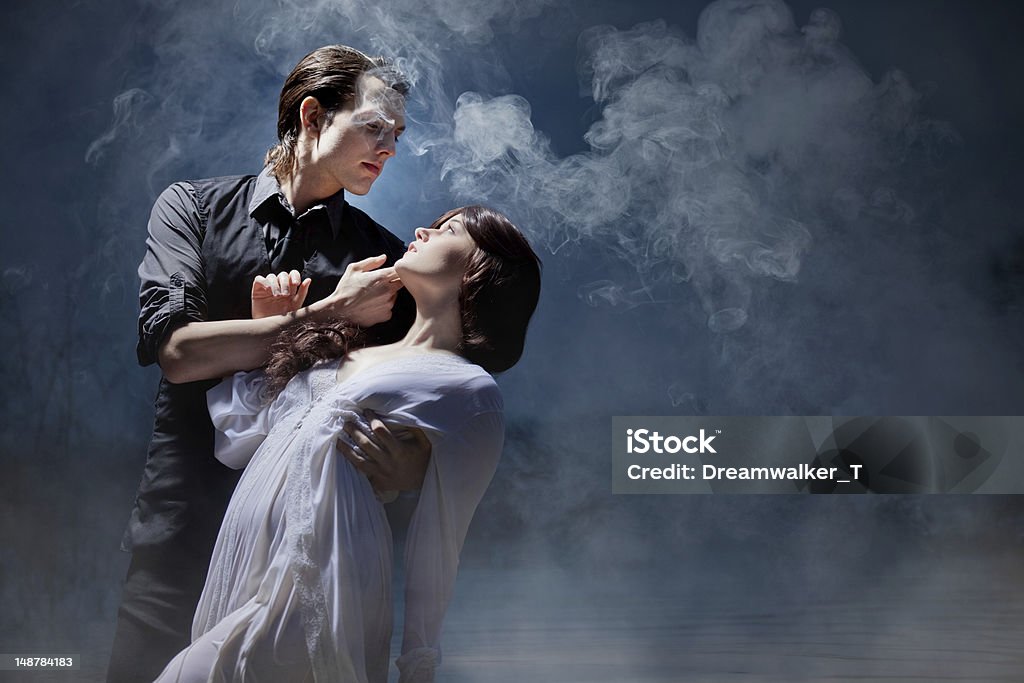 Love Between Dark and Light Romantic, dramatic image of seductive dark-haired man dressed in black holding beautiful young woman in classical white dress against a misty, midnight setting. Fainting Stock Photo
