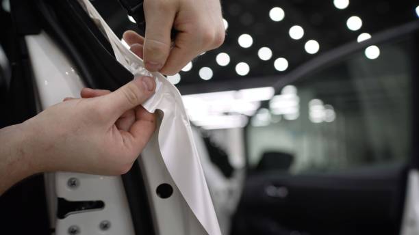 Car detailing. Man sticking protective film on a car body at the vehicle service station, focused on the car front. Concept of car body protection with special films. A worker sticks a white film on a car. stock photo