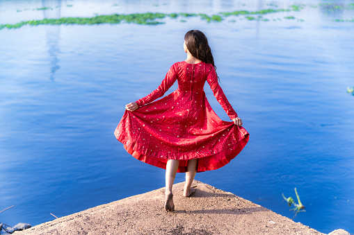 Full body back view of young female holding up polka dot red dress with one hand while raising another standing on wooden pier near blue lake