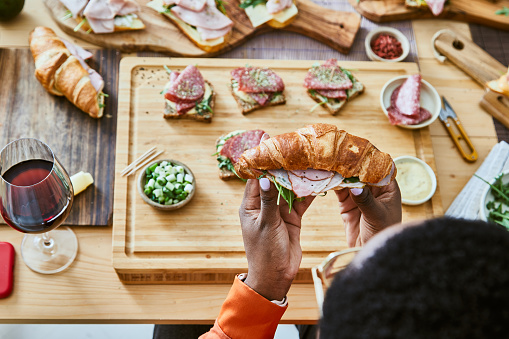 Beautiful young girl preparing sandwiches for social gathering or birthday event, with fresh green salad, ham, cheese, mayo, mustard, tomato, cucumber, pesto, cottage cheese, cream cheese, avocado, Spanish onion with healthy seasoning, herbs and spices, served on a wooden home table, representing a wellbeing and a healthy lifestyle, food indulgence and joy, an image with a copy space