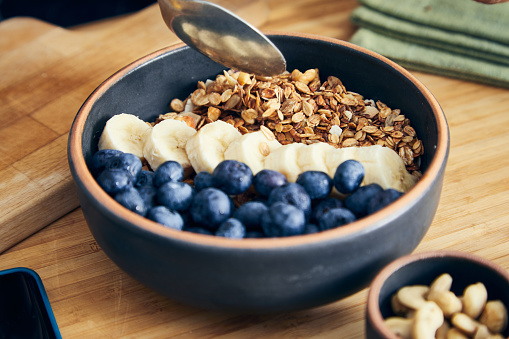 A healthy oatmeal for breakfast, with fresh organic fruit, banana and blueberry, nuts and grains, at a wooden table in a modern white kitchen, representing a healthy lifestyle and wellbeing, an image with a copy space