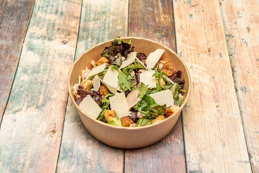 Caesar salad with parmesan cheese flakes, croutons and lettuce shoots with arugula in cardboard bowl for home delivery on wooden table
