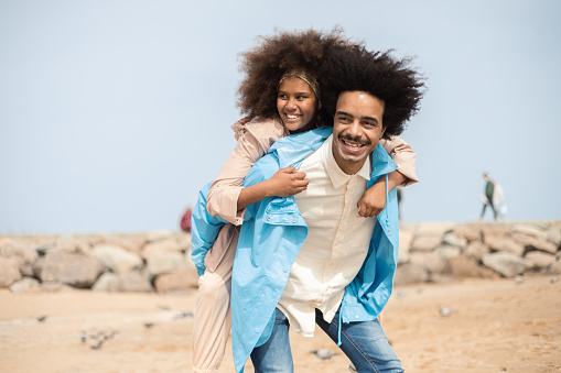 Portrait of a happy African American man carrying his teenage daughter on back and enjoying on the beach