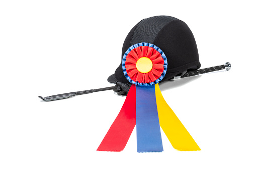 A multi-colored equine Champion award ribbon clipped a helmet, whip and gloves sitting on a white background.