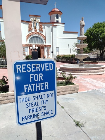 Humorous sign on not stealing a priest's parking space in a church parking lot. Funny sign on being non sinful it terms of parking lot behavior, found in Las Cruces NM. Our Lady of Good Health church blurred in background.