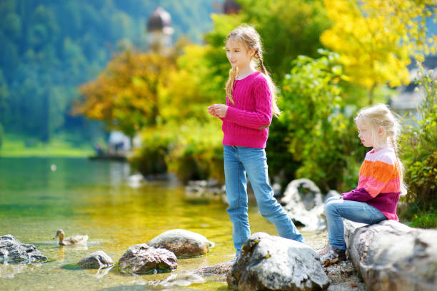 очаровательные сестры, играющие на озере кенигсе в германии в теплый летний день. симпатичные дети весело кормят уток и бросают камни в озе� - konigsee стоковые фото и изображения