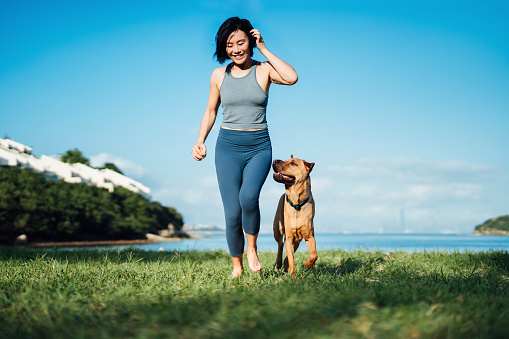 Happy young Asian woman and her pet dog running by the beach against clear blue sky, playing and enjoying time together in the nature outdoors. Living with a dog. Love and bonding with pet