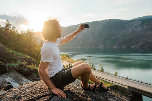 Man with curly hair in white t-shirt making selfie on the background of the fjord in Norway