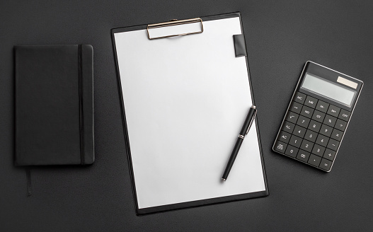 Clipboard with blank sheet of paper, notepad and calculator on the black desk. Top view. Copy space.