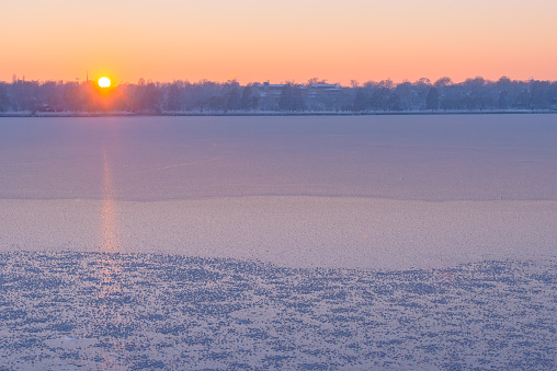 Scenic view of a frozen lake in the snowy city park, with frosted trees in the background in beautiful orange sunset light. Extreme weather, polar vortex, freezing temperature, winter alert concept.