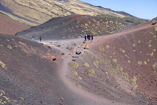 Mount Etna, Sicily, Italy - April 27, 2023: People walking the path of the slopes of Crater Silvestri of volcano Mount Etna on the sunny day. Stones and solidified lava, withered vegetation