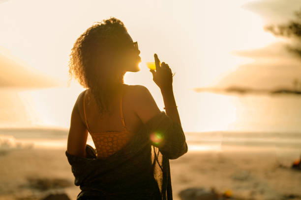 열대 해변에 좌초할 때 칵테일 주스를 마시는 아프리카 여성. 노란 비키니를 입고 자연의 아름다움을 만끽하는 젊은 여행자. 방랑벽과 여행 개념. - luxury women chaise longue fashion 뉴스 사진 이미지